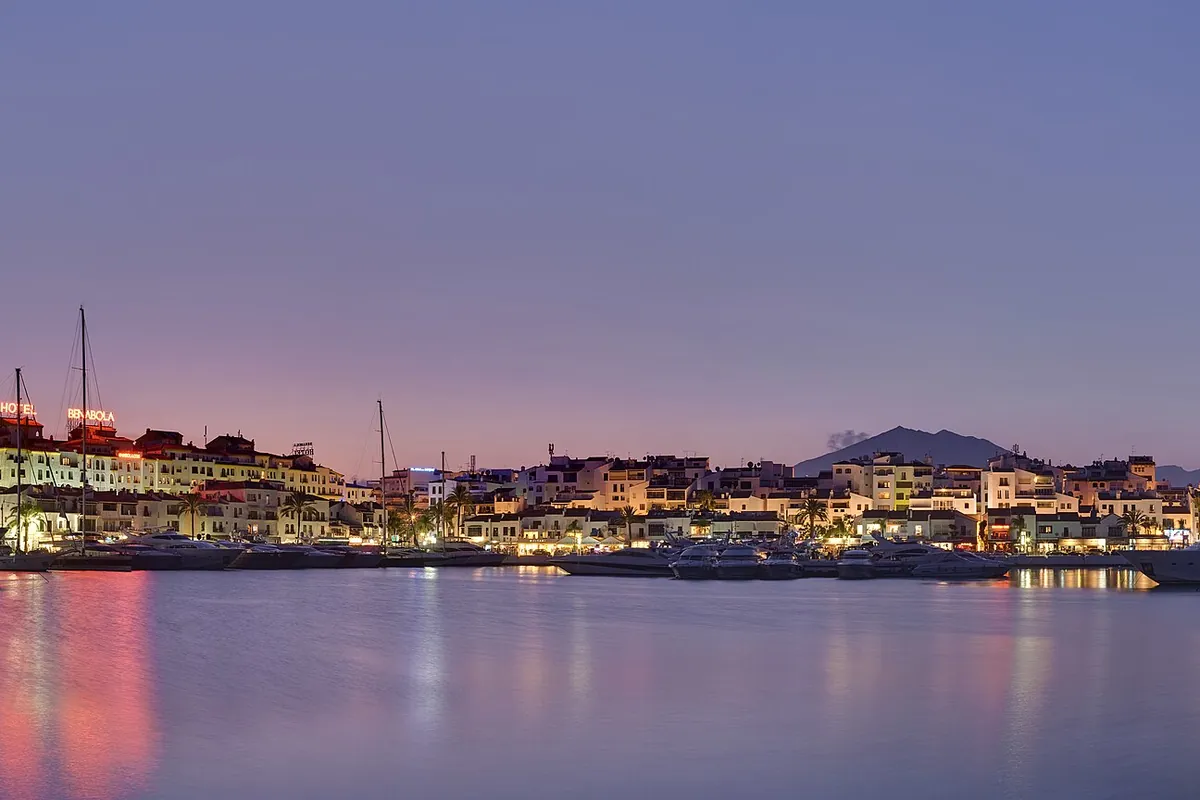 A marina at dusk with lit buildings along the quay, yachts moored at the water's edge and a violet sky reflected in calm water
