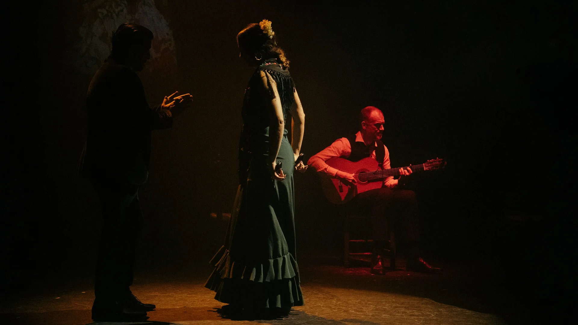 A flamenco dancer in a dark green ruffled dress stands in warm stage light beside a seated guitarist and a singer in silhouette on a dim stage