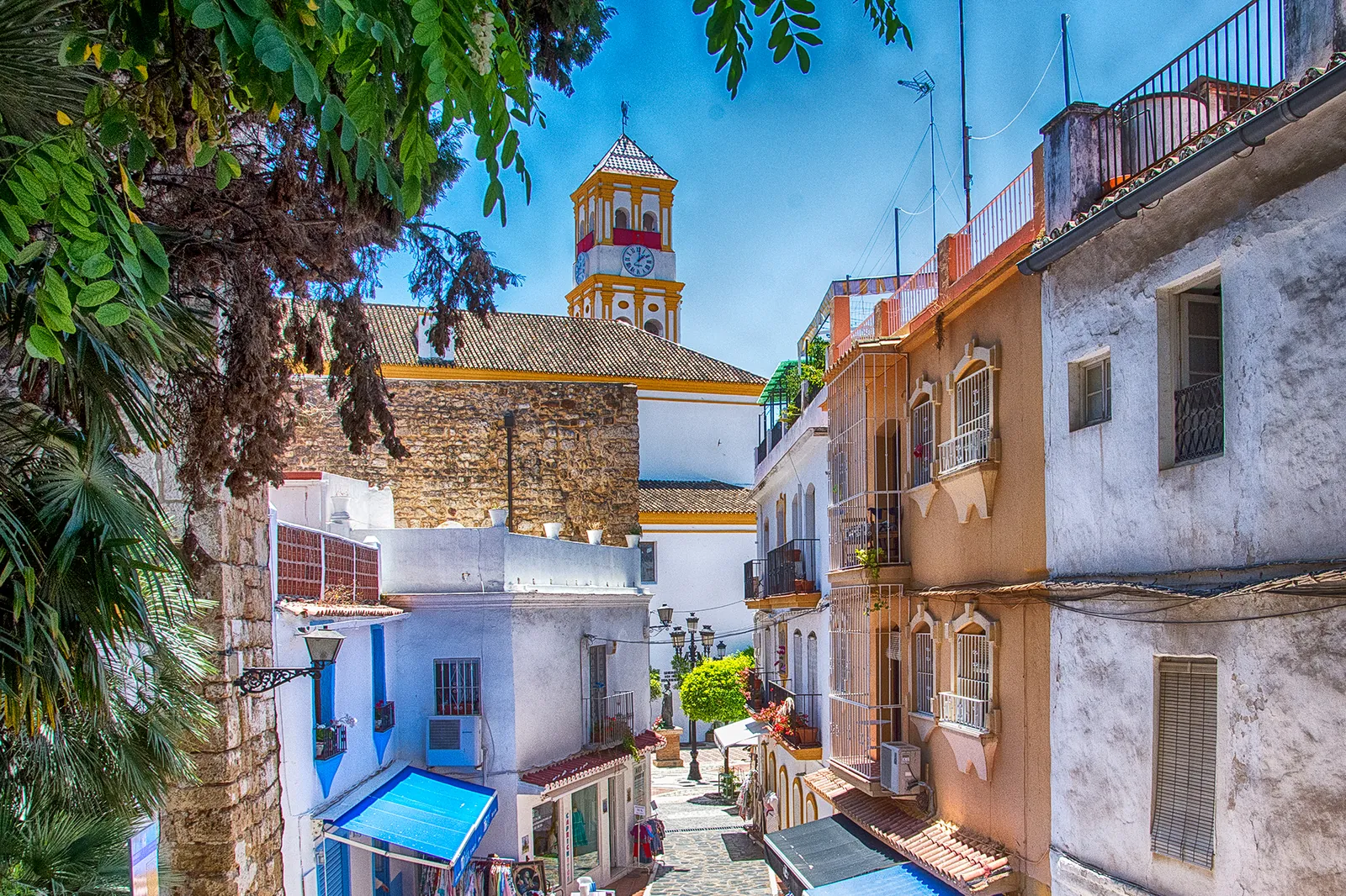 A steep lane in Marbella's old town with colourful houses, a blue awning and a church bell tower in the distance