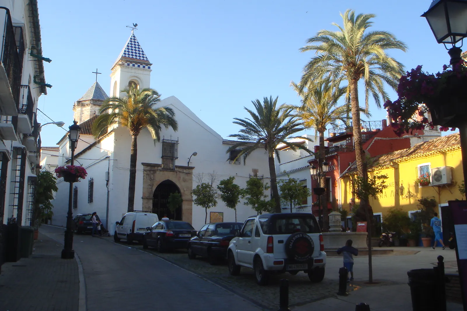 A whitewashed church with a small tower on a quiet square in Marbella, with palm trees, parked cars and a yellow house beside it